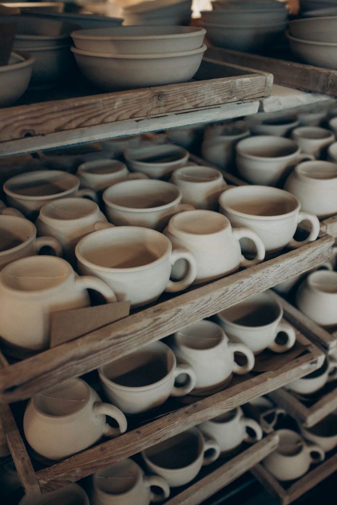 Rows of handmade ceramic cups and bowls on rustic wooden shelves in a pottery studio.